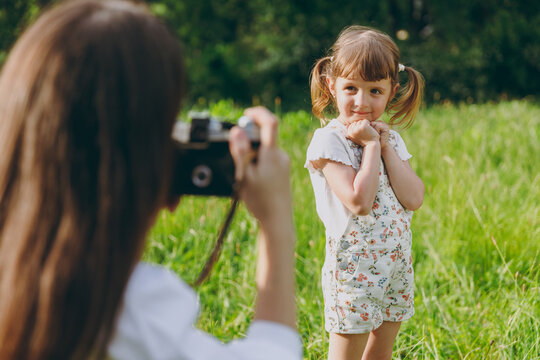 Photographer woman in white clothes take picture shot on photo camera of cute caucasian child baby girl 5-6 years old on green grass Mommy rest with little kid daughter outdoor. Love family concept