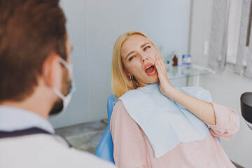 Young suffering sick woman look at dentist sit at office chair indoor light cabinet hold cheeck visiting stomatologist with toothache painful problem wait cure Healthcare oral enamel caries treatment.