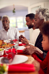 Multi Generation Family Hold Hands Around Table At Home Saying Grace Before Eating Christmas Meal