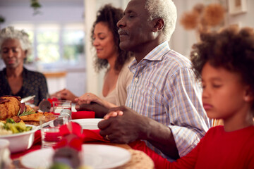 Multi Generation Family Hold Hands Around Table At Home Saying Grace Before Eating Christmas Meal