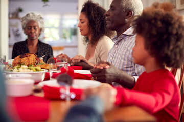 Multi Generation Family Hold Hands Around Table At Home Saying Grace Before Eating Christmas Meal