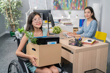 Disabled women with glasses and Asian Korean beauty sits in wheelchair at corporate desk on lap holding a cardboard box of packed accessories, changing positions, getting promoted