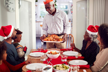 Grandfather Carrying Turkey As Multi-Generation Family Sit Down To Eat Christmas Meal Together