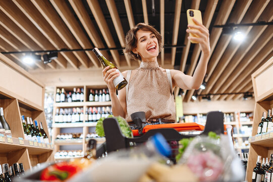Young fun woman in casual clothes shopping at supermaket store with grocery cart hold white wine alcohol hold bottle do selfie shot on mobile phone inside hypermarket Purchasing gastronomy concept