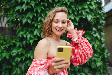 Young smiling satisfied happy fun blonde woman wear pink dress using mobile cell phone typing message chat standing outdoor on green ivy leaves background. People urban summer time lifestyle concept