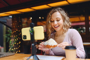 Young smiling happy woman wear casual clothes sit at table in cafe shop eat breakfast take picture on mobile cell phone do selfie shot photo of croissant drink coffee relaxing in restaurant indoors