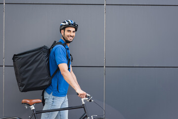 Cheerful muslim deliveryman with backpack and bike walking near building outdoors