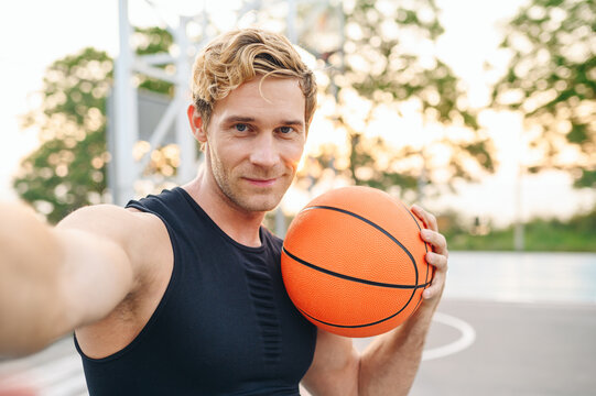 Close up muscular young sporty sportsman man in black sports clothes training doing selfie shot pov on mobile phone hold ball play at basketball game playground court Outdoor courtyard sport concept