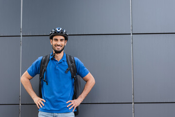 Smiling arabian courier with helmet and backpack standing near building outdoors