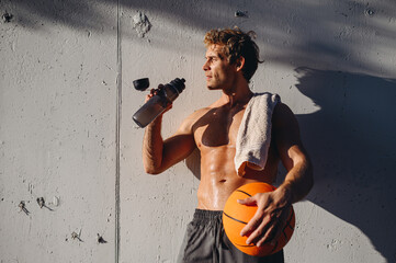 Young serious minded sportive sunlit sportsman man with naked torso training hold in hand basket...