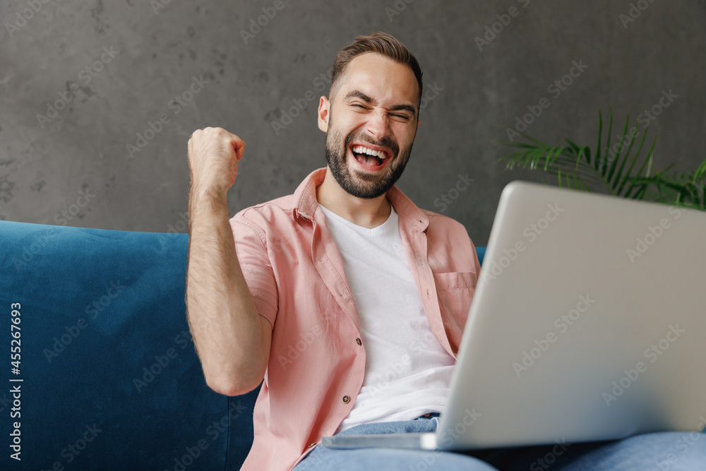 Canvas Prints Young satisfied fun happy smiling smart man 20s wear casual clothes hold use work on laptop pc computer do winner gesture clench fist sitting on blue sofa at home flat indoors rest relax on weekends