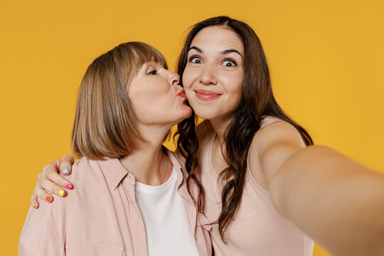Close up two young smiling daughter mother together couple women in casual beige clothes doing selfie shot pov on mobile phone kissing hug isolated on plain yellow color background studio portrait