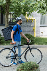 Fototapeta premium Side view of positive arabian courier with thermo backpack standing near bike outdoors
