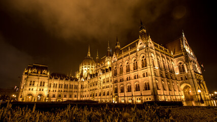 Fototapeta na wymiar Parlement de Budapest illuminé la nuit, photo prise à ras du sol sous un ciel nuageux