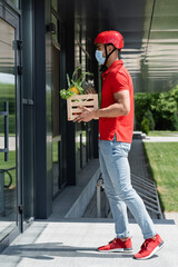 Side view of arabian courier in protective helmet and medical mask holding vegetables near building