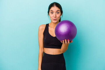 Young caucasian fitness woman holding a ball isolated on blue background shrugs shoulders and open eyes confused.