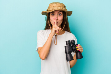 Young caucasian explorer woman holding binoculars isolated on blue background keeping a secret or...