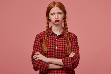 indoor shot of young redhead ginger female, wears checkered black red shirt, keeps her hands crossed, looks into camera with sad, negative facial expression, curves her lips.