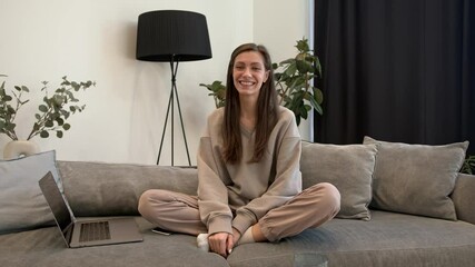 Attractive brunette woman, smiling, wearing beige home clothes sitting on a couch and waving hello on the camera. Sofa with laptop and mobile phone, gimbal shot