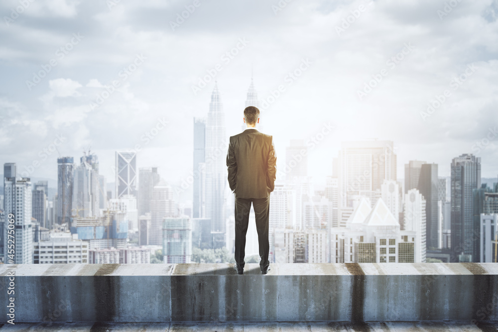 Poster Back view of young businessman standing on concrete edge of building on city background with mock up place in sky. Success, future, vision and leadership concept.