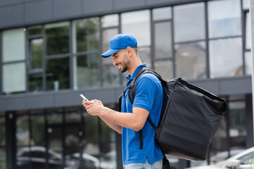 Side view of cheerful arabian courier with thermo backpack using smartphone on urban street