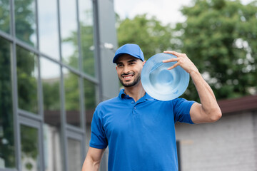 Cheerful arabian deliveryman in cap holding bottle of water outdoors