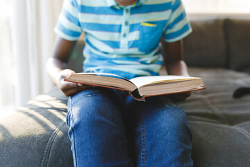 Midsection of african american boy reading book and sitting on couch in living room