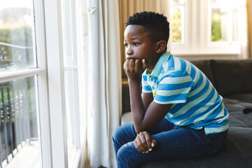 Thoughtful african american boy sitting and looking out of window in living room