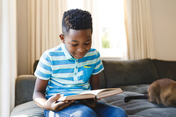 Smiling african american boy reading book and sitting on couch with cat in living room - Powered by Adobe