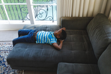 Thoughtful african american boy lying on couch looking out of window in living room