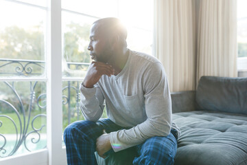 Worried, sad african american man thinking, sitting on couch looking out of window in living room - Powered by Adobe