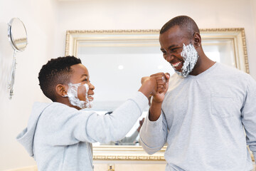 African american father with son having fun with shaving foam, bumping fists in bathroom