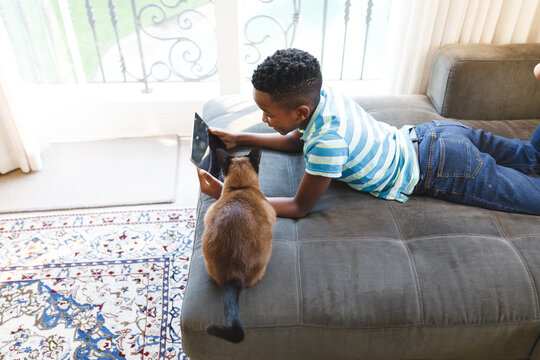 African american boy using tablet and lying on couch with cat in living room