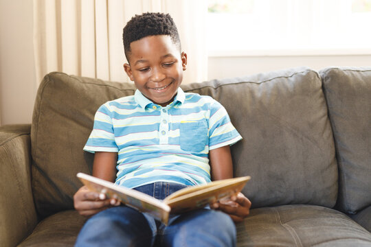 Smiling african american boy reading book and sitting on couch in living room