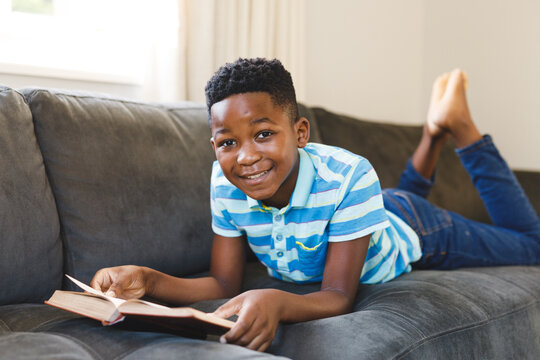 Portrait of smiling african american boy reading book and lying on couch in living room - Powered by Adobe