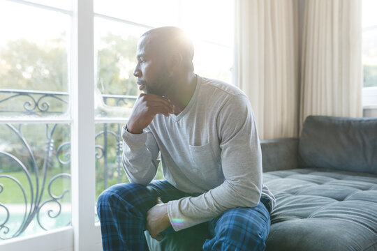 Worried, sad african american man thinking, sitting on couch looking out of window in living room