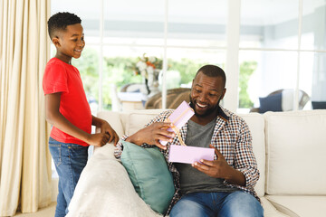 African american father opening gift from his son and smiling in living room