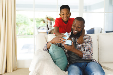 African american father opening gift from his son and smiling in living room - Powered by Adobe