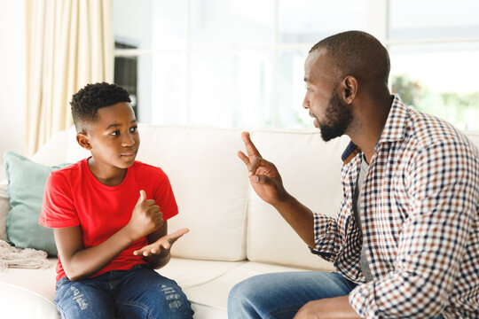 Happy african american father with son sitting on couch in living room talking sign language