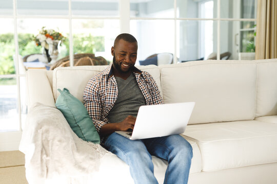 Smiling african american man using laptop and sitting on couch in living room