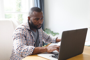 African american man sitting at table in dining room, working remotely using laptop
