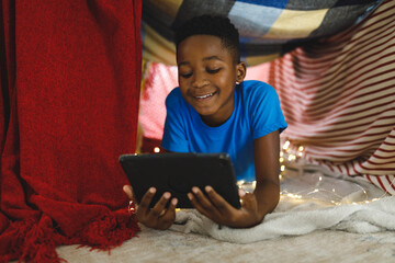 Happy african american boy lying in blanket fort, using tablet