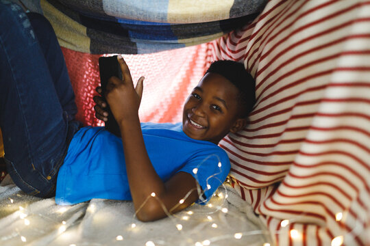 Portrait Of Happy African American Boy Sitting In Blanket Fort, Using Tablet