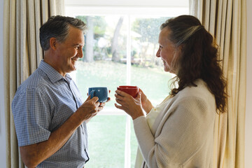 Happy senior caucasian couple standing next to window, holding cups