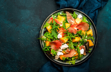 Fresh salad with cantaloupe melon, prosciutto, soft cheese and arugula on blue background, top view, copy space
