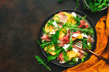 Salad with melon, jamon, goat cheese and arugula on brown background, top view, copy space