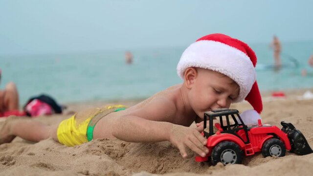 Happy boy in a santa claus hat plays with a toy car on the beach. A child in Christmas is resting on the sea. Copy space