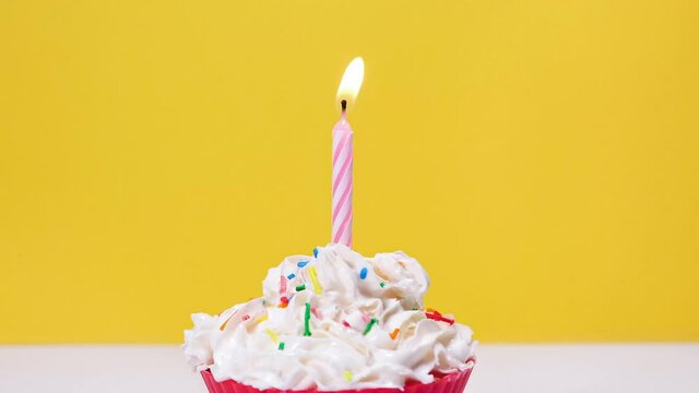 Close up view on tasty capcake with whipped cream and one burning cake candle isolated on yellow background. Blowing out one cake candle. Full HD slow motion happy birthday or anniversary video.