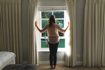 Fototapeta premium Thoughtful senior caucasian woman in bedroom, standing next to window, opening curtains