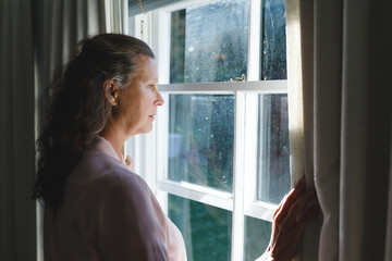 Thoughtful senior caucasian woman in bedroom, standing next to window, widening curtains